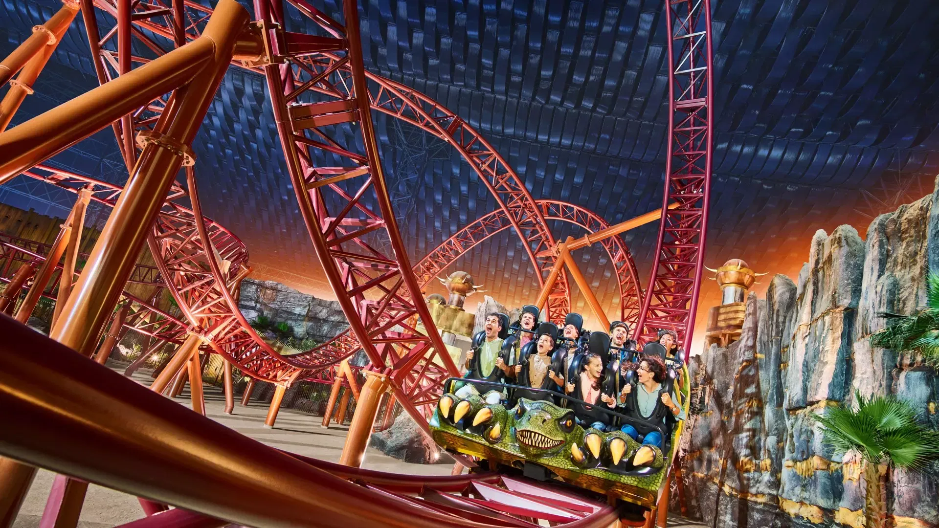 A group of people ride a red roller coaster through loops beneath an indoor ceiling at IMG Worlds of Adventure Theme Park.