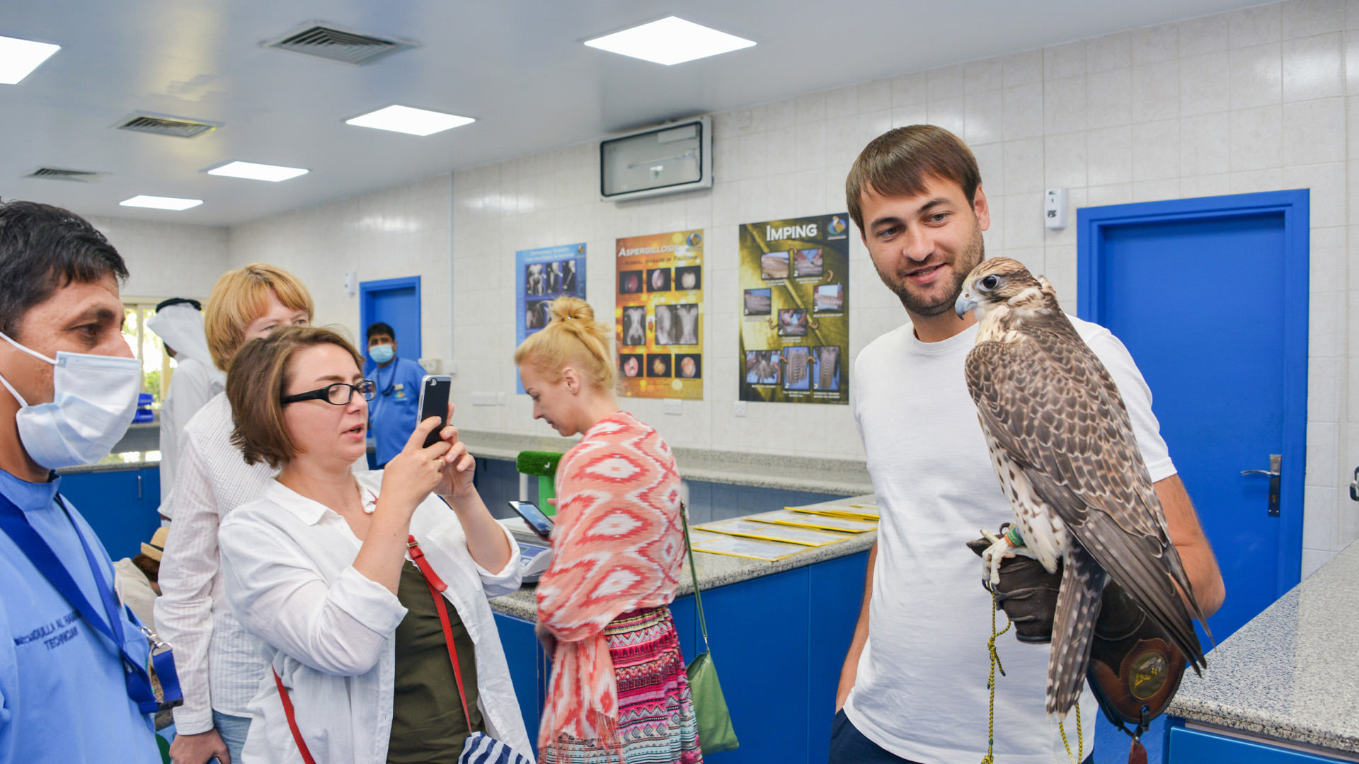 Visitors tour a bright clinical room while a man holds a falcon on his gloved hand and a woman photographs them at Abu Dhabi Falcon Hospital.
