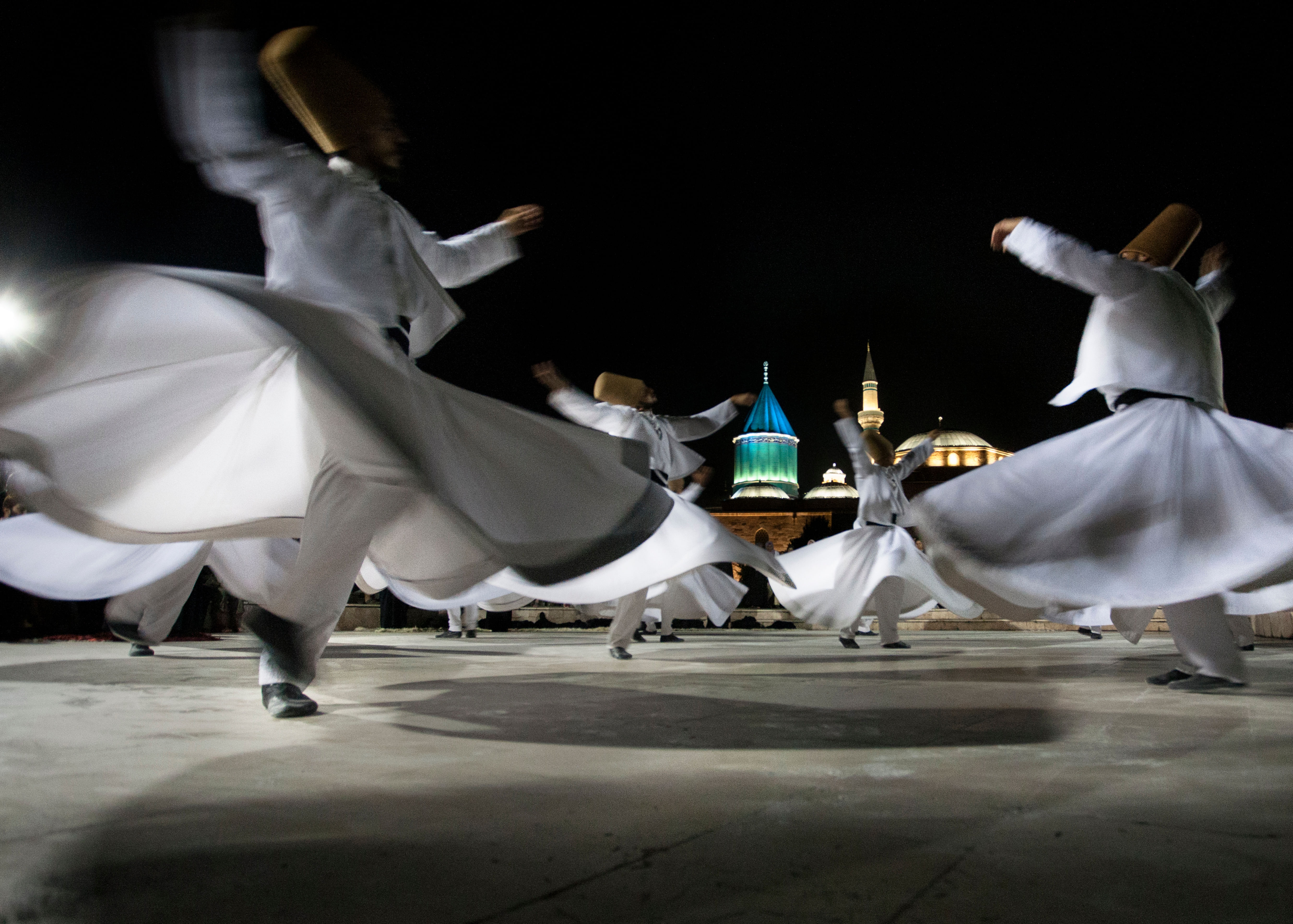 “Whirling Dervishes dressed in traditional white robes and tall hats, spinning gracefully during the Sema ceremony in a dimly lit room.”