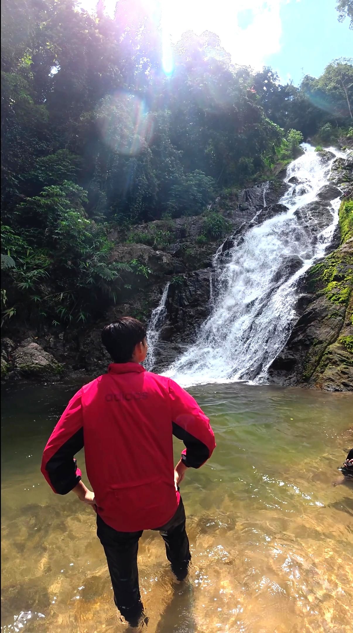 A person stands in shallow water looking up at the beautiful Jeram Perlus waterfall cascading down rocks in a lush Malaysian rainforest.