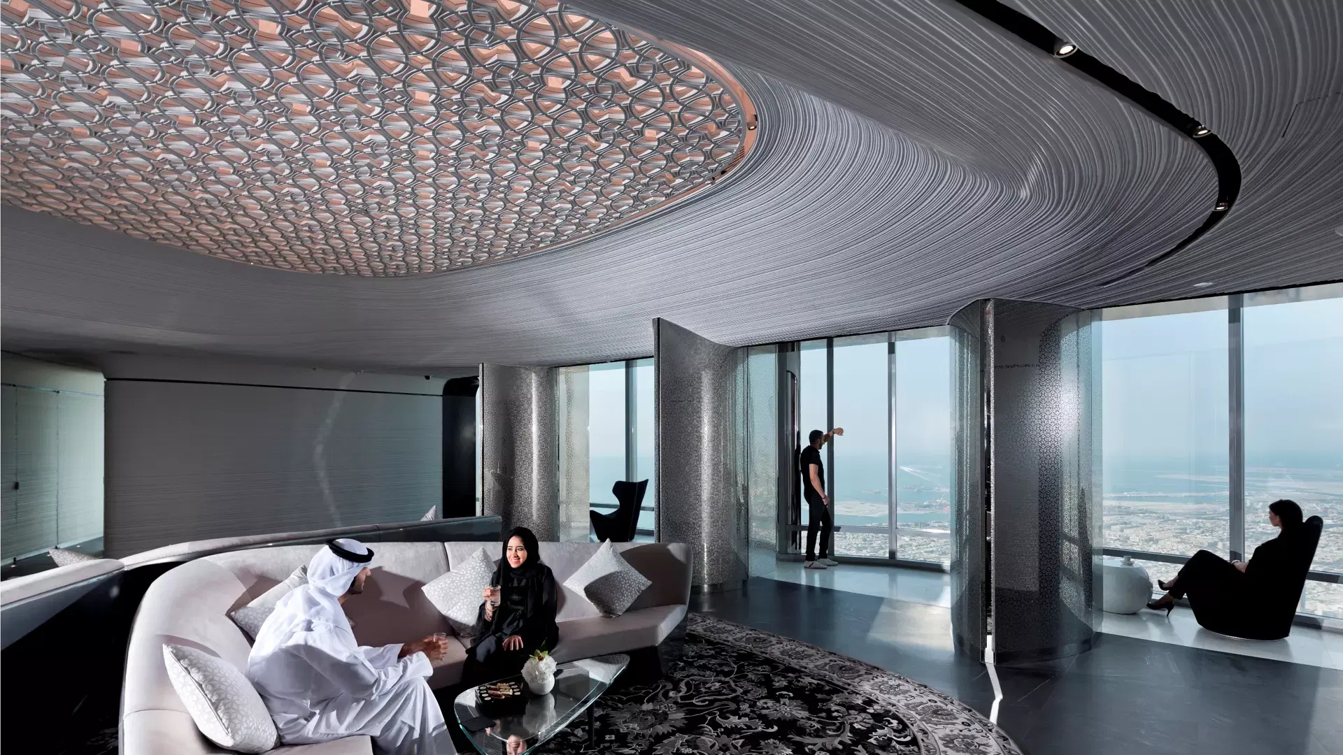 Guests seated on a curved sofa beneath a patterned metal ceiling in a lounge high in the tower, another visitor looking out to the sea at Burj Khalifa