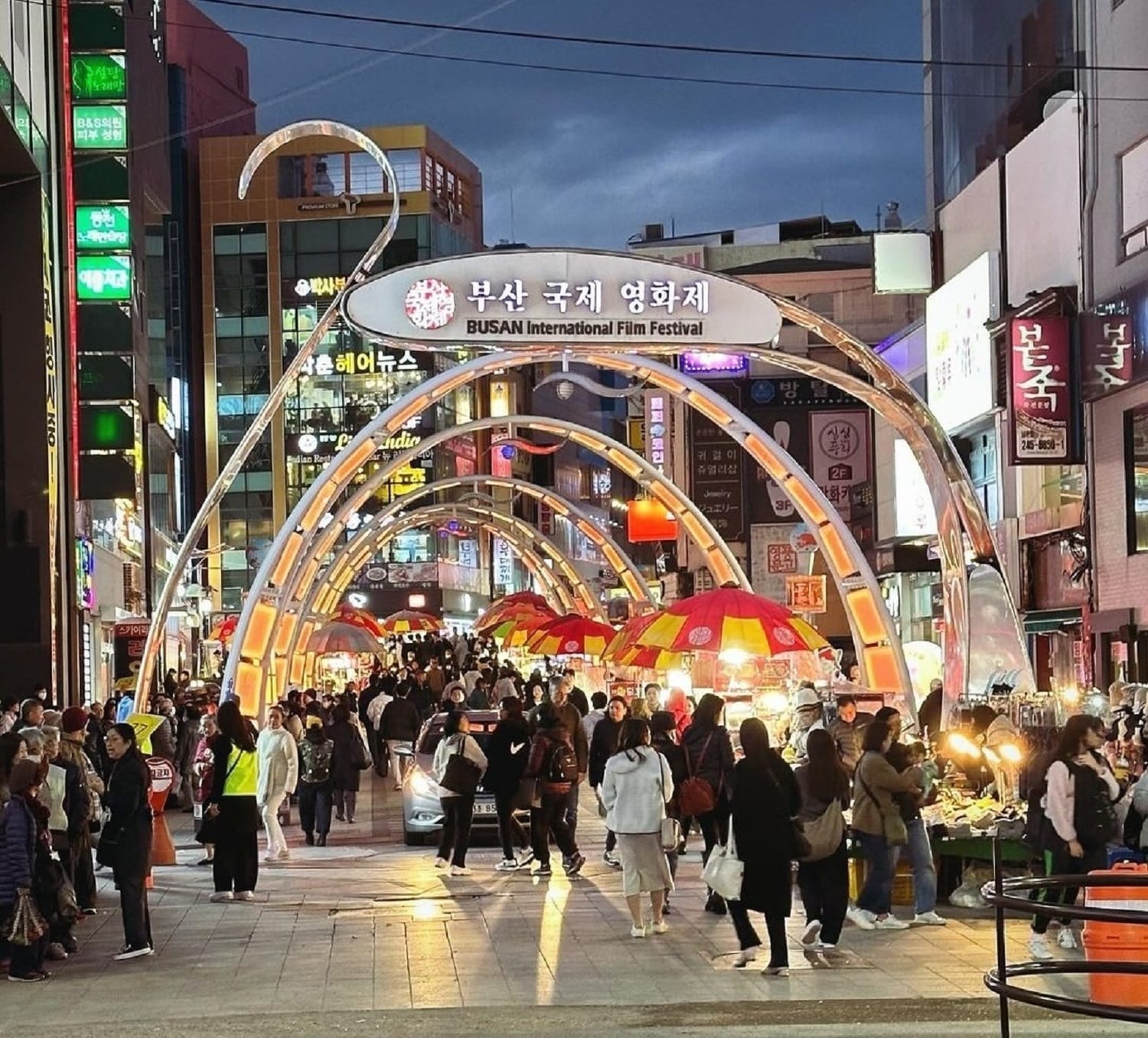 Crowds walk beneath illuminated arches on a lively street in BIFF Square, Busan, decorated for the film festival at night.
