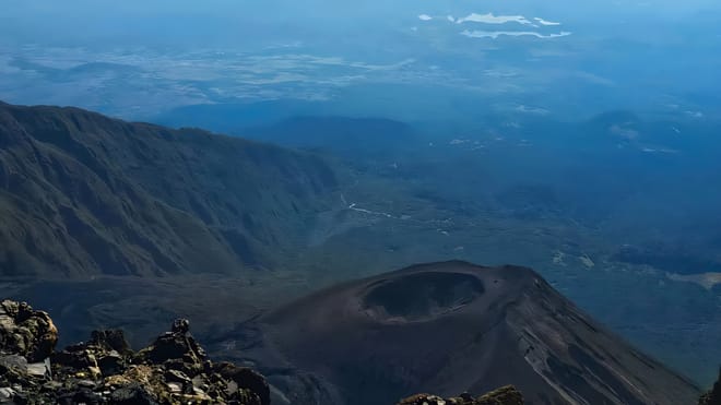 High-angle view looking down into the volcanic crater of Mount Meru with surrounding slopes and distant clouds.