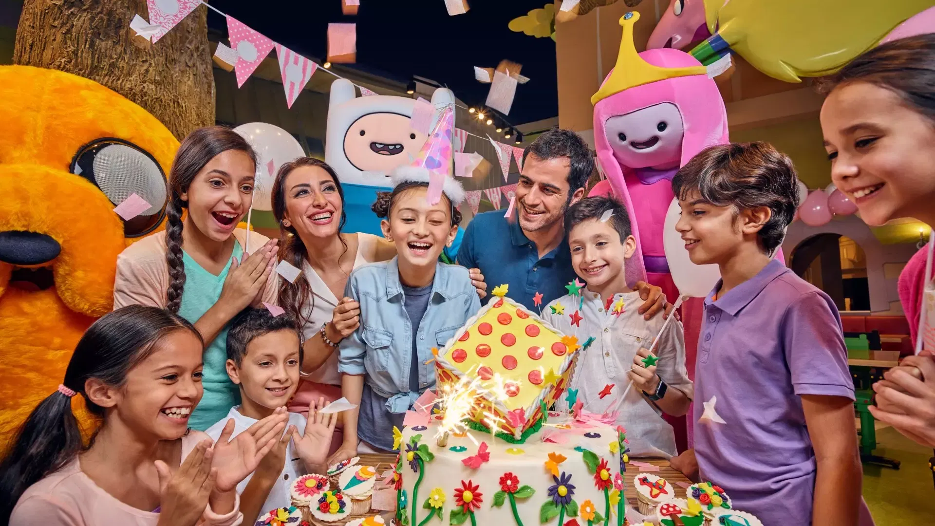A diverse group of smiling children and adults gather around a brightly decorated birthday cake with lit sparklers, surrounded by colorful cartoon characters and party decorations at IMG Worlds of Adventure Theme Park.