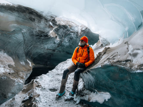 man in orange jacket sitting in the natural ice cave with the blue hue of the ice