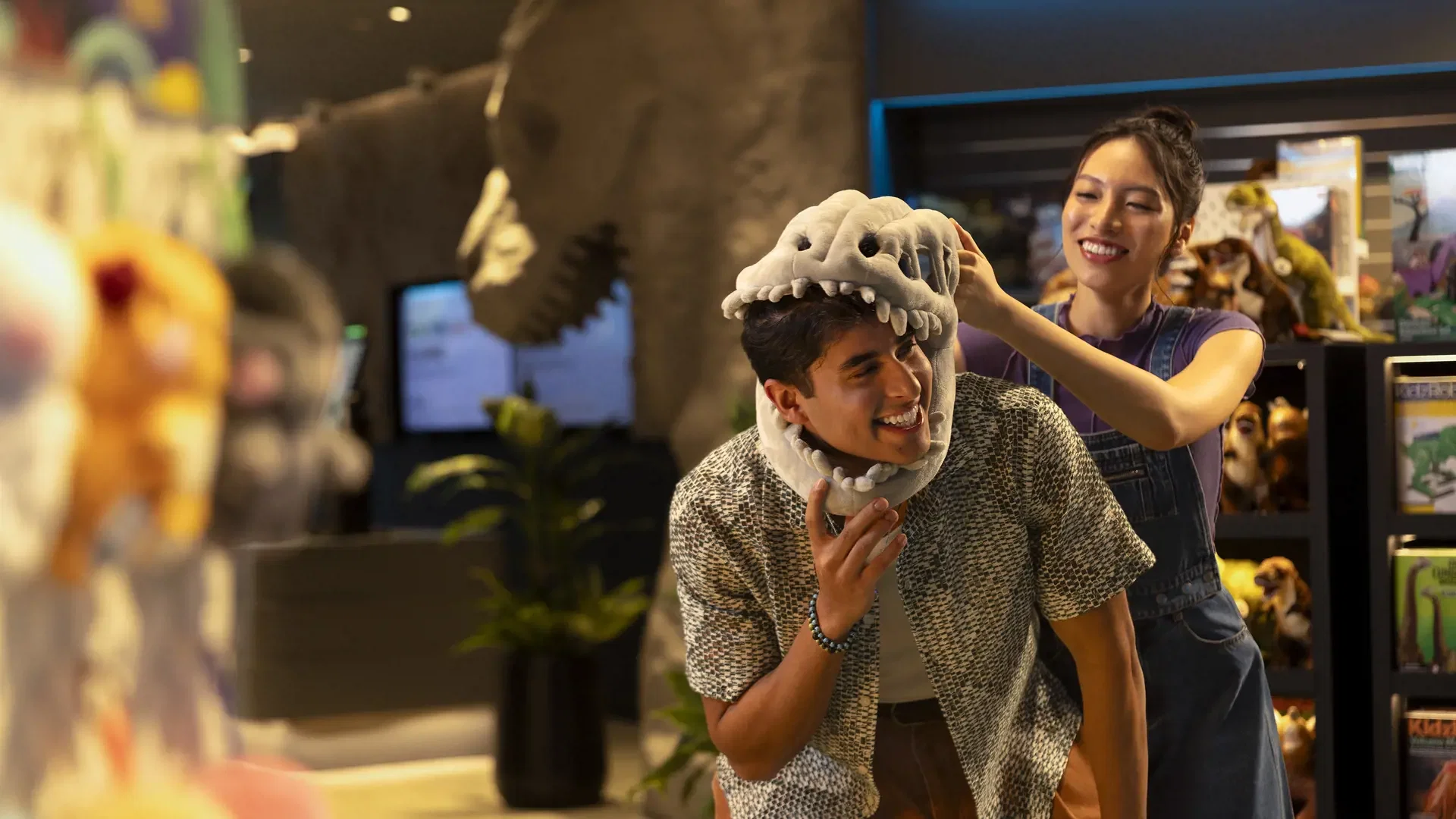 A smiling woman playfully places a plush dinosaur skull hat on a laughing man inside a souvenir shop at Exploria in Mandai Singapore Ticket.