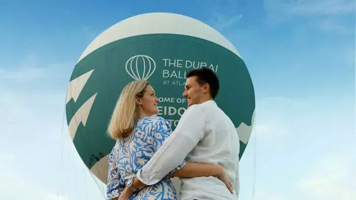 A couple embraces in front of a large branded balloon under a bright blue sky at The Dubai Balloon at Atlantis.