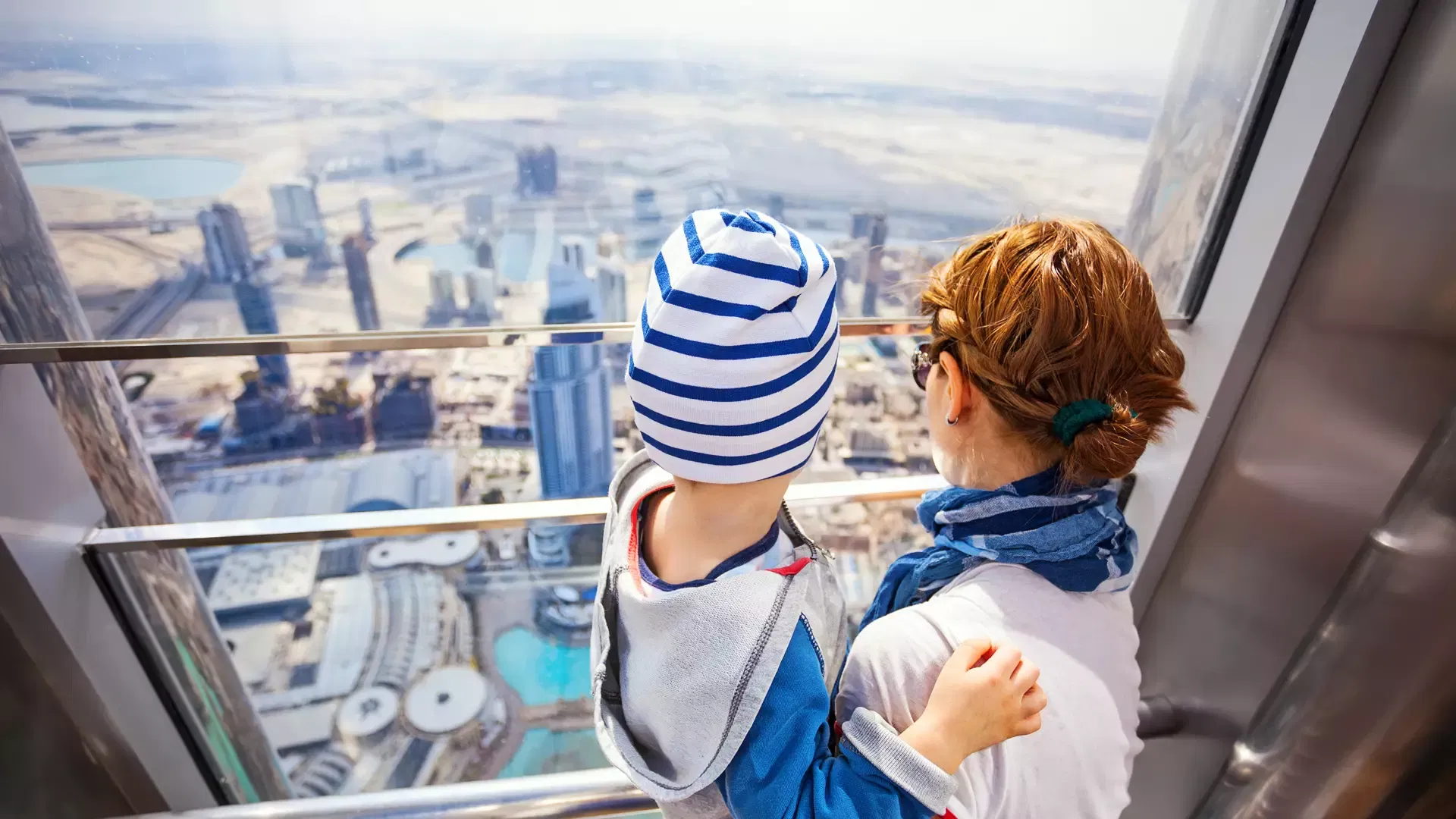 A woman holding a small child in a blue striped hat as they look down through the glass at the city far below at Burj Khalifa