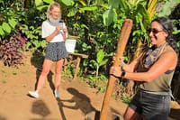 Two female tourists smiling while using a large wooden mortar and pestle to crush coffee beans in a lush garden.