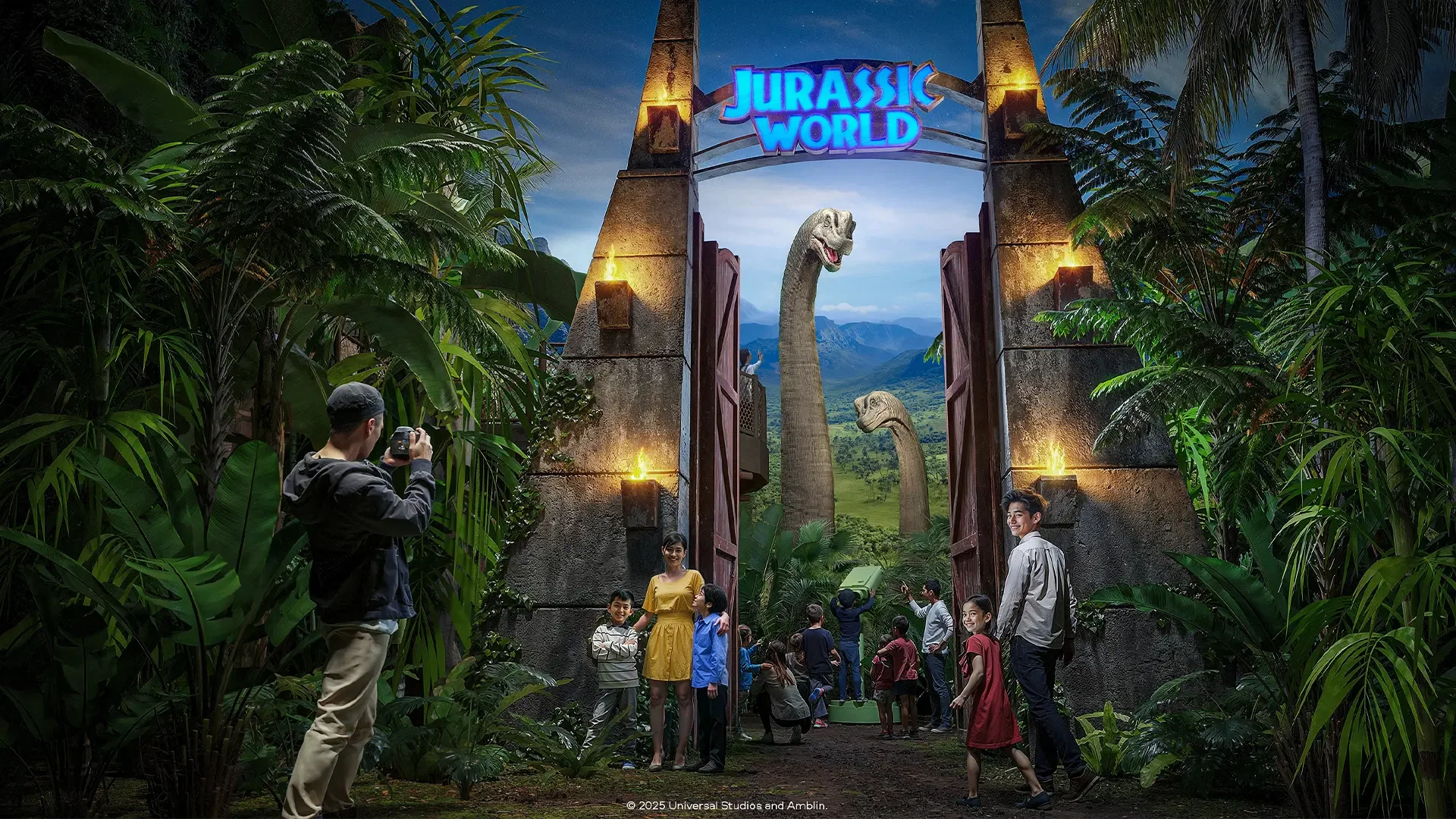 A group of visitors stands beneath the large park entrance gate while a photographer in the foreground takes a picture of the scene at Jurassic World: The Experience Bangkok Ticket.