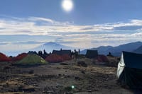 Several colorful trekking tents pitched on a rocky mountain plateau with a large peak and bright sun in the background.