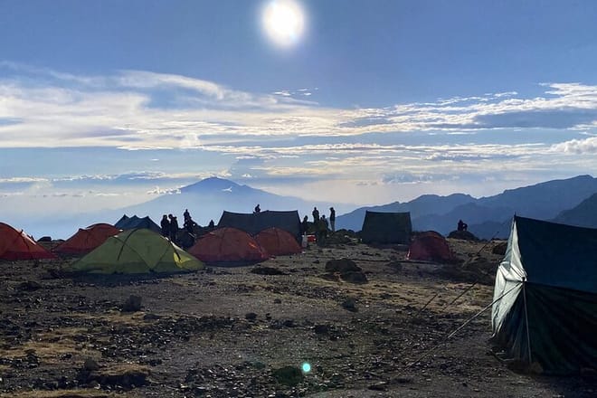 Several colorful trekking tents pitched on a rocky mountain plateau with a large peak and bright sun in the background.