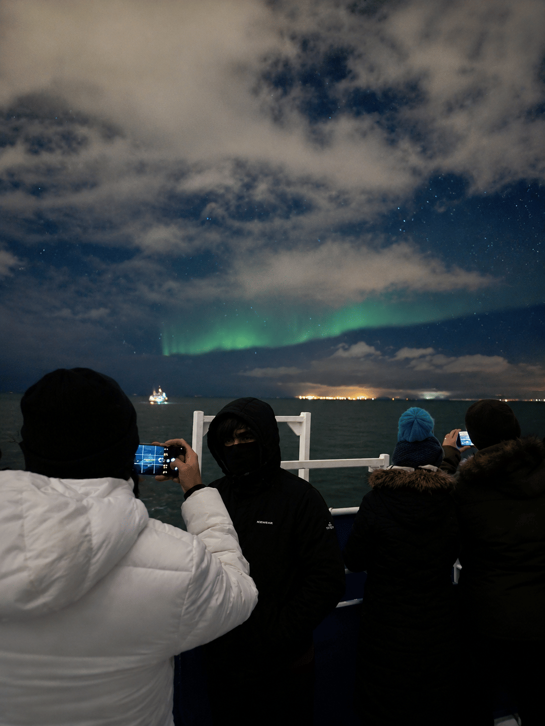 Passengers photographing the Northern Lights from a boat deck beneath a cloudy night sky.
