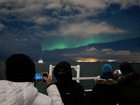 Passengers photographing the Northern Lights from a boat deck beneath a cloudy night sky.
