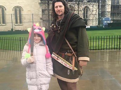 A viking guide stood with a young girl, who is posing with the Viking's seax knife, in the rain. They are next to York Minster.