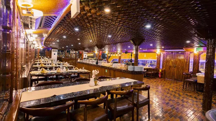 Interior cabin of a boat featuring long dining tables with chairs, a wooden ceiling, and ambient lighting at Alexandra Dhow Cruise in Dubai Marina.