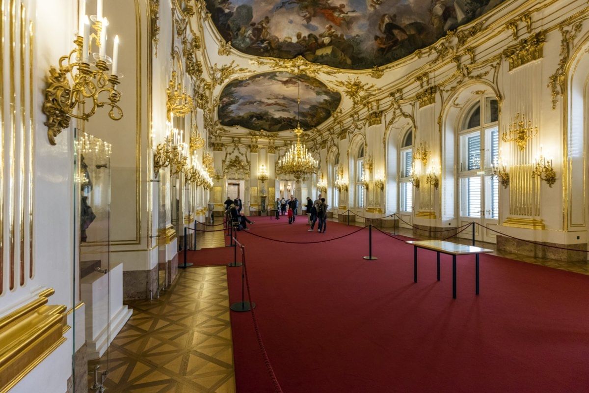 Opulent hall inside Schönbrunn Palace featuring crystal chandeliers, gilded walls, detailed ceiling paintings, and visitors walking along a 