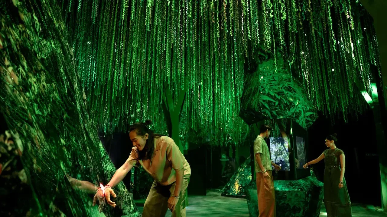 Several people explore a dimly lit room featuring hanging green foliage and glowing tree-like structures at Exploria in Mandai Singapore Ticket.