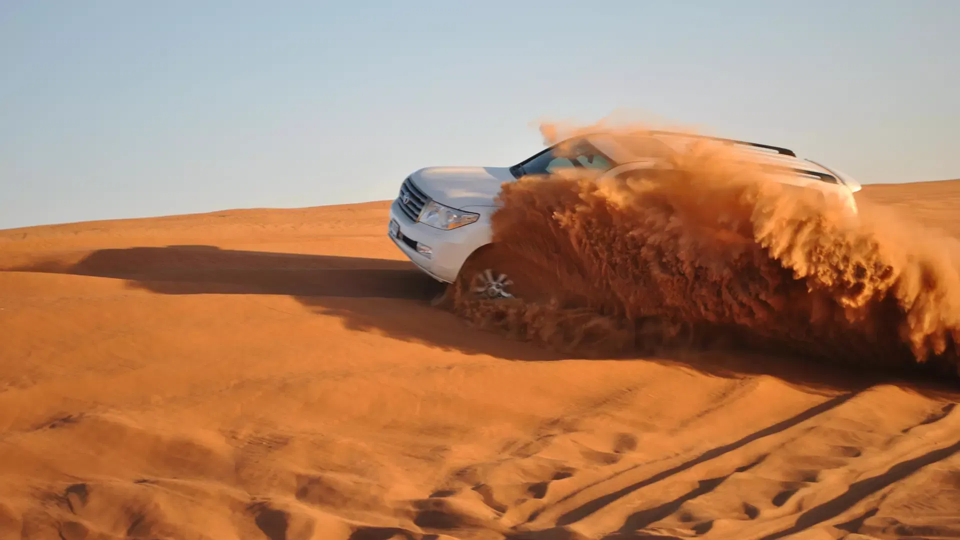 A white SUV kicks up a thick spray of reddish-brown sand while driving across rolling dunes at Desert Safari + Al Faqaa Camp.