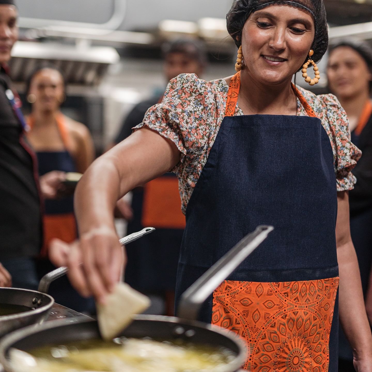A smiling woman in a floral blouse, a blue and orange patterned apron, and a hairnet is gently placing a samoosa into a pan of oil. 