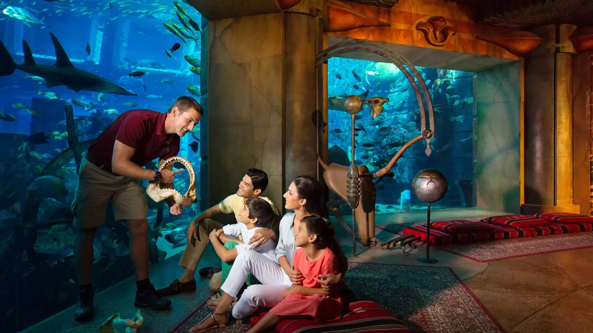 A guide holding a shark jawbone talking to a seated family in front of the floor-to-ceiling aquarium windows of the Lost World Aquarium at Aquaventure Waterpark