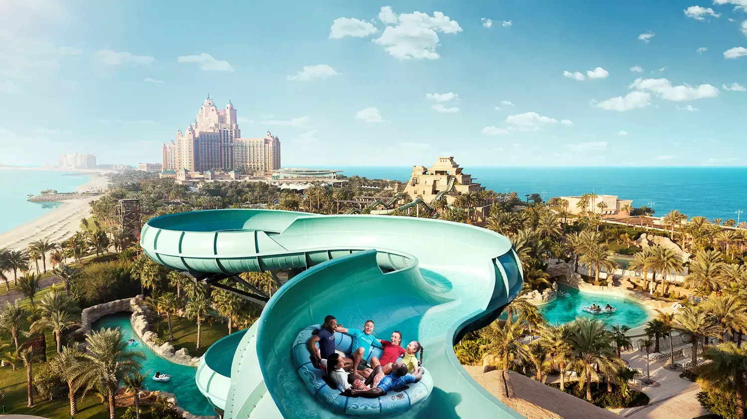A group of riders in a large round raft sweeping through a curved green flume high above the palm gardens, with the Atlantis The Palm hotel and the beach behind, at Aquaventure Waterpark