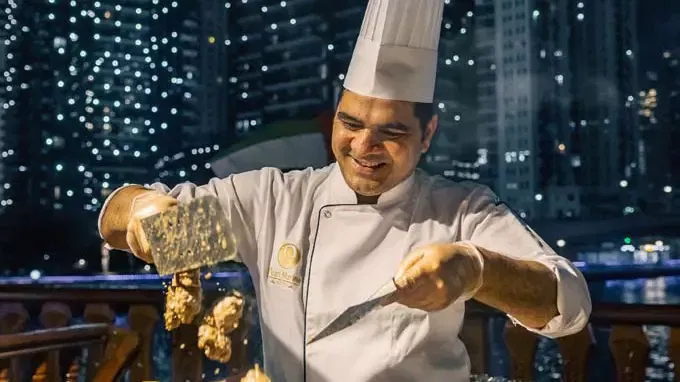 A chef in a white uniform and tall hat smiles while using spatulas to flip food against a backdrop of illuminated city buildings at Alexandra Dhow Cruise in Dubai Marina.