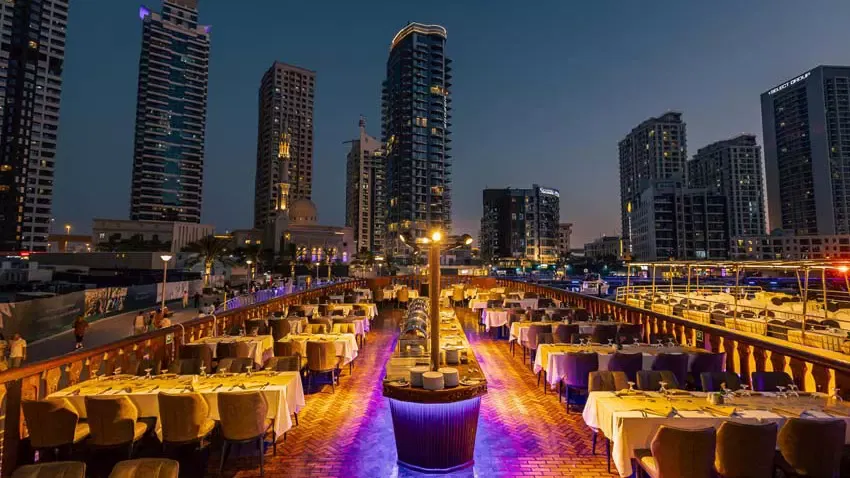 A wide angle shot of a rooftop dining area on a boat showcases neatly set tables and city skyline views at Alexandra Dhow Cruise in Dubai Marina.