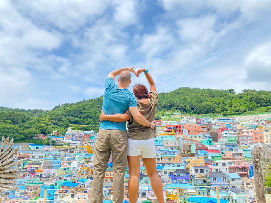 A couple standing with colorful hillside houses of Gamcheon Culture Village in the background.