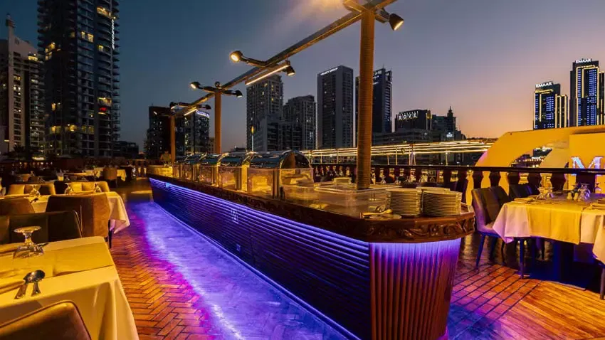 An outdoor dining deck on a boat features a buffet station with purple accent lighting and surrounding tables at Alexandra Dhow Cruise in Dubai Marina.