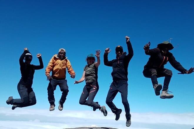 A group of four trekkers jumping for joy in the air on the summit of Mount Kilimanjaro against a clear blue sky.