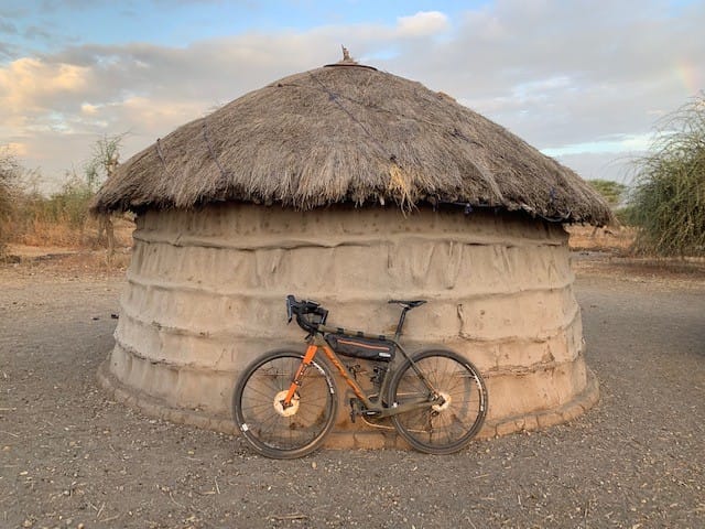 An orange touring bicycle parked in front of a traditional circular Maasai mud hut with a thatched roof outdoors.