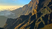 Steep, rocky mountain cliffs glowing in warm sunset light above a sea of white clouds.