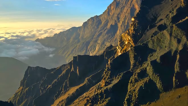 Steep, rocky mountain cliffs glowing in warm sunset light above a sea of white clouds.