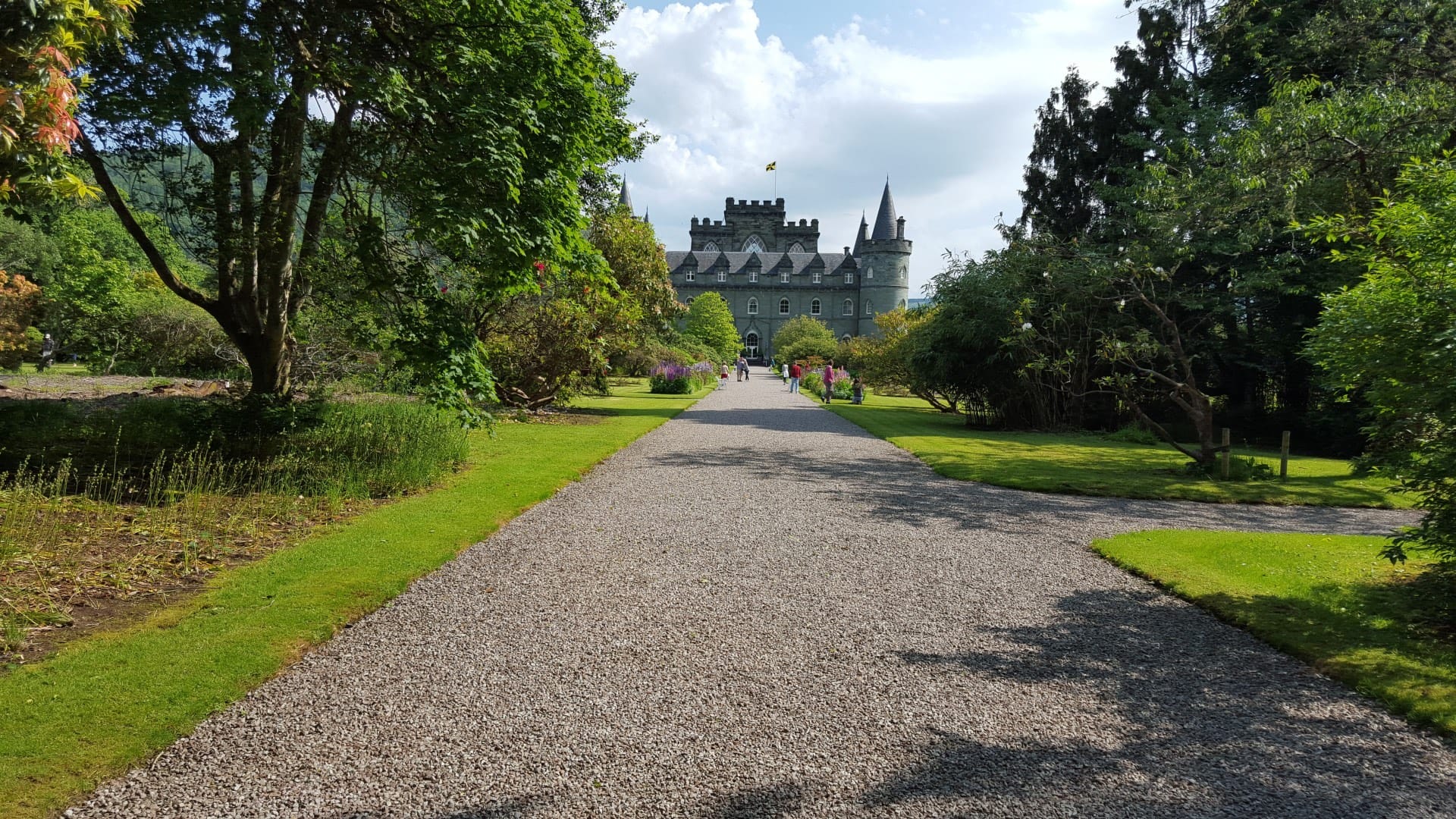 A grand, stately castle with grey stone walls and conical turrets stands amidst lush green gardens. The castle, Inveraray Castle, is framed