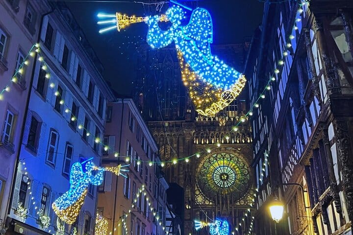 Rosace illuminée de la Cathédrale de Strasbourg entre deux bâtiments à colombages, encadrée par des anges lumineux bleus.