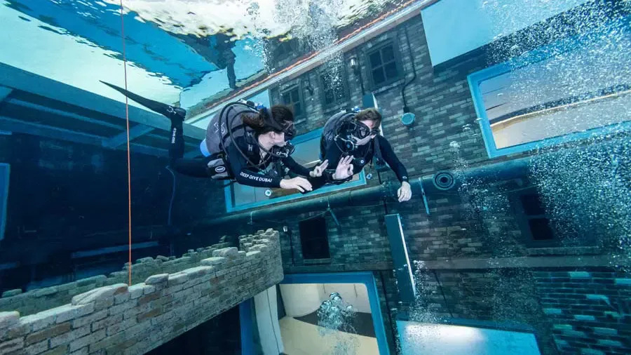 Two scuba divers navigate past a submerged brick wall with windows and pipes at Deep Dive Dubai.