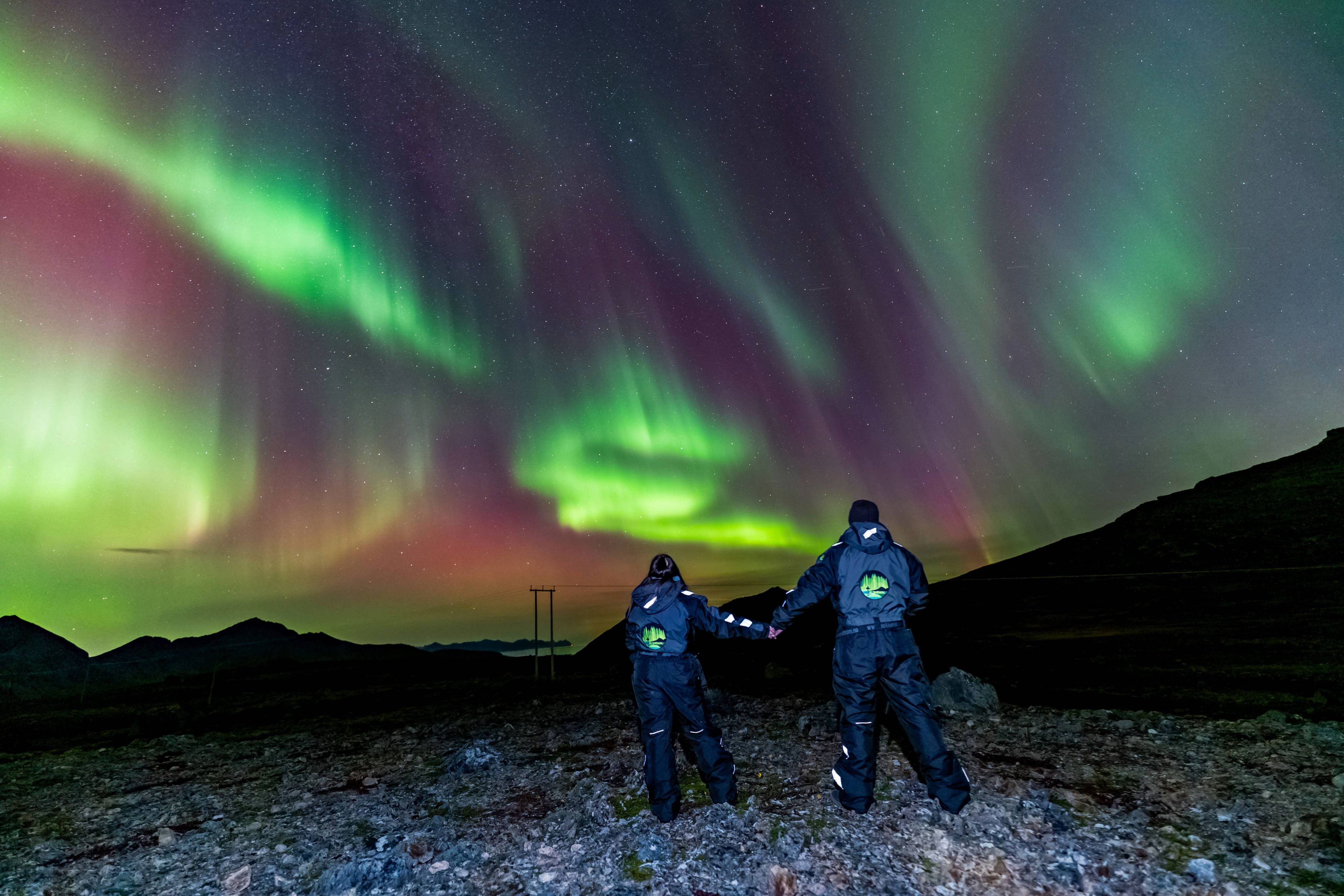 Couple holding hands on rocky ground under a vibrant pink and green Northern Lights display during a Tromsø small-group tour.