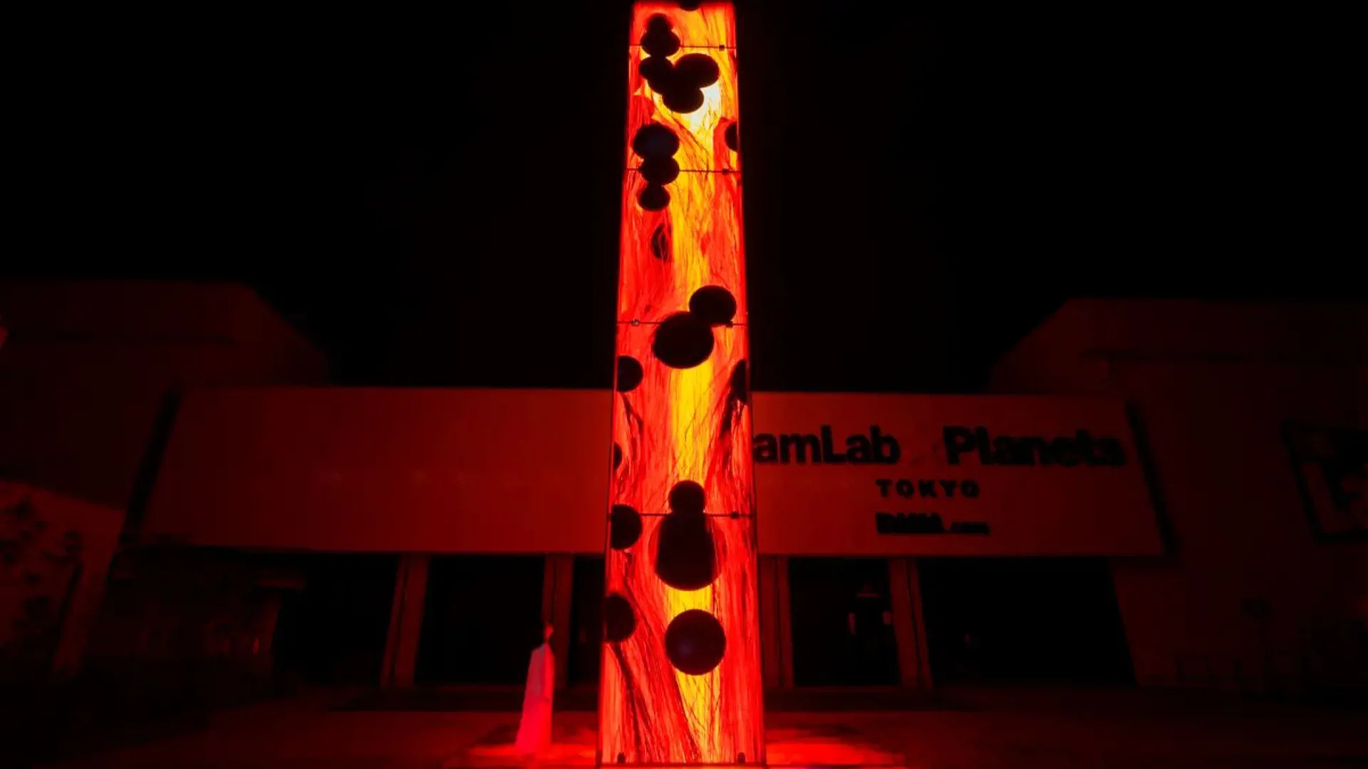 A person stands beside a tall, glowing red column displaying abstract black bubble patterns outside a dark building at COMBO: Tokyo Skytree + teamLab Planets TOKYO Tickets.