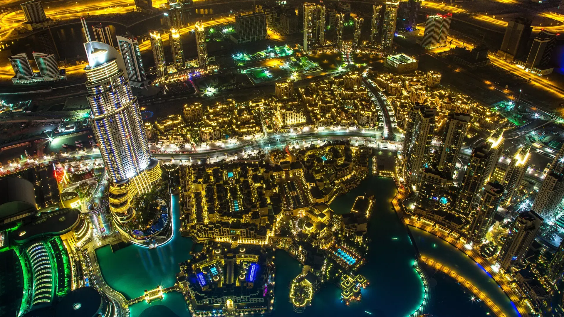 Night view straight down over Downtown Dubai, the lit tower blocks, curving roads and turquoise lake glowing gold below, from Burj Khalifa