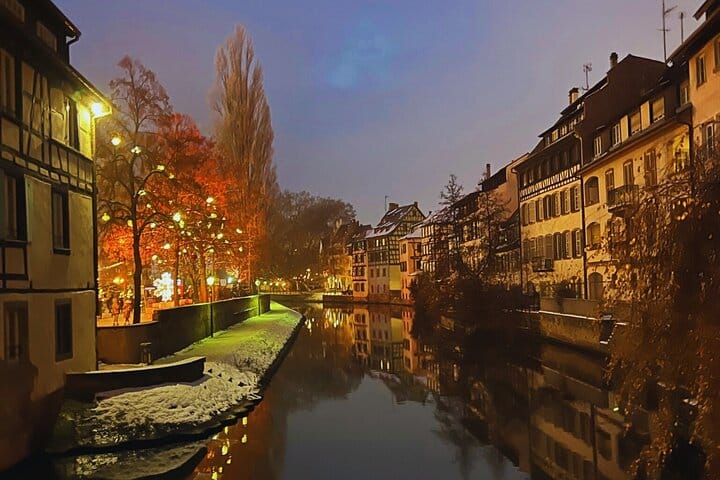 La Petite France of Strasbourg at night, with illuminated half-timbered houses alongside a canal, trees decorated with twinkling lights.