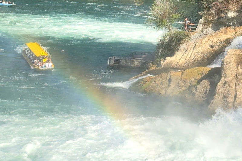 As Cataratas do Reno com o arco-íris sobre a água e o barco que leva até a rocha no meio da queda.