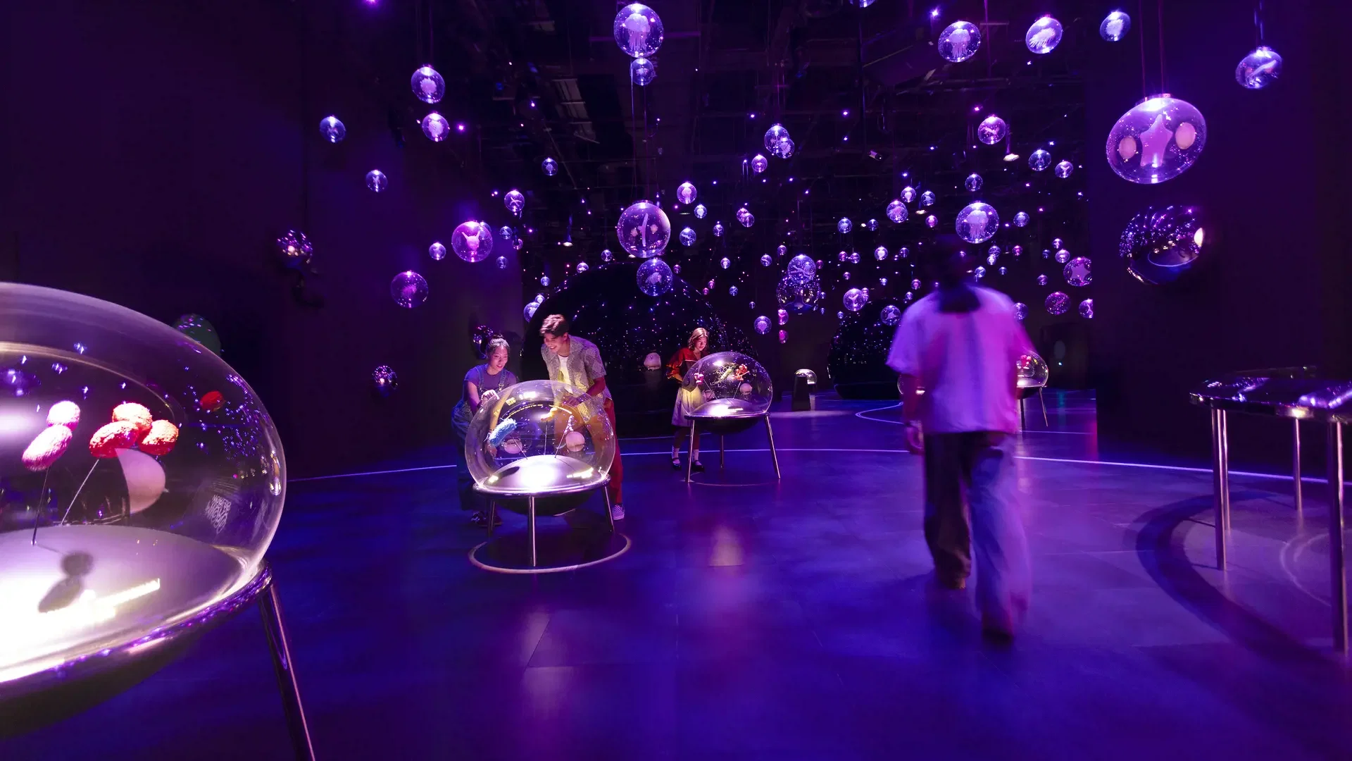 Visitors walk through a dark, purple-lit exhibition space filled with suspended glowing globes and tabletop interactive exhibits at Exploria in Mandai Singapore Ticket.