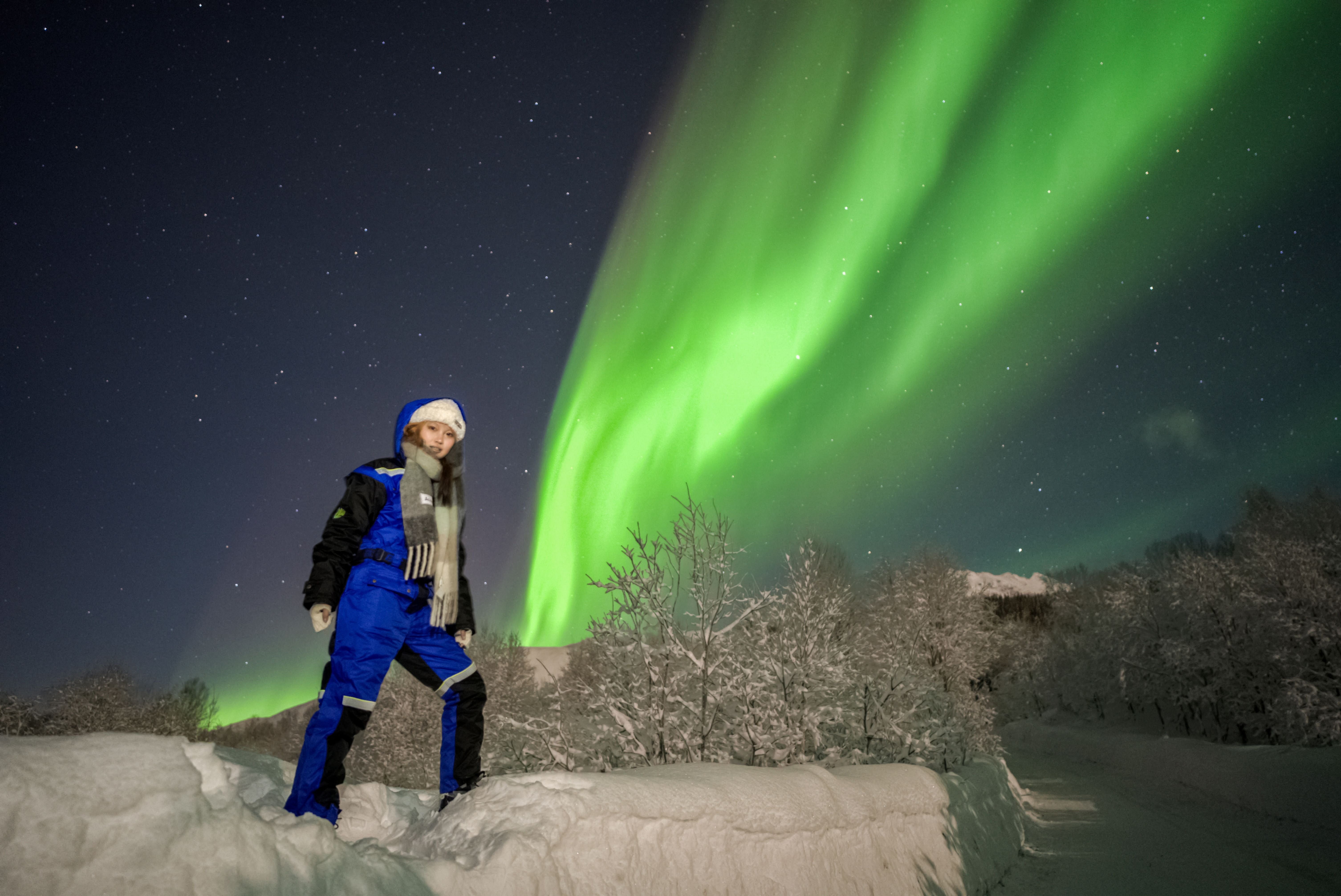 Traveler wearing a thick blue thermal suit standing on snow under a vibrant green Northern Lights display in Tromsø.