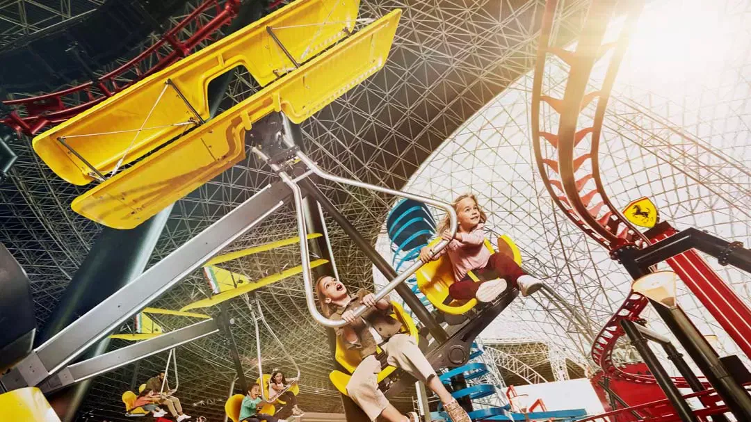 A woman and a young girl swinging on a yellow aeroplane-themed ride beneath the glass roof, with red coaster track curving beside them, at Ferrari World Abu Dhabi