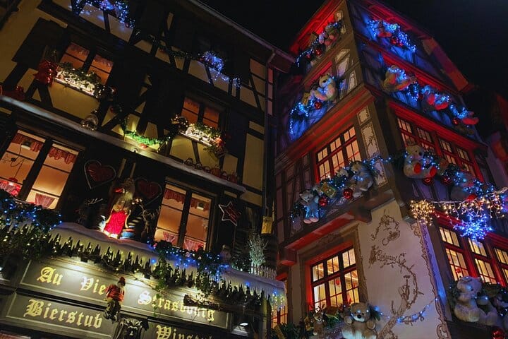 Half-timbered house façades on Rue du Maroquin illuminated with blue garlands and decorated with teddy bears at night.