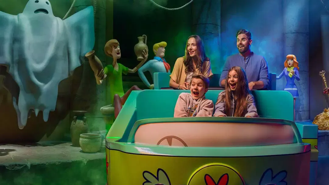 Four people ride in a green vehicle past a glowing ghost and animated characters in a dark room at Warner Bros. World Abu Dhabi Theme Park.