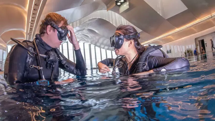 Two scuba divers wearing masks and wetsuits float in a shallow indoor pool while talking to each other near a large windowed hall at Deep Dive Dubai.