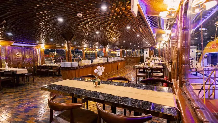 An interior shot shows a dining hall with a patterned wooden ceiling, marble-topped tables set with runners, and a long buffet counter along the back wall at Alexandra Dhow Cruise in Dubai Marina.