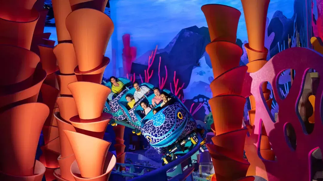 A roller coaster car full of laughing passengers navigating an indoor dark ride surrounded by giant, brightly colored coral reef formations at Sea World Abu Dhabi.